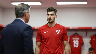 A 21-year-old Croatian central defender in a solid red jersey, standing in a dressing room with a determined expression...