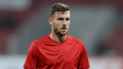 A mid-30s anonymous KV Mechelen player, waist-up, focused expression, wearing a solid red jersey, blurred stadium...