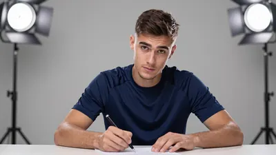 A 22-year-old male centre-back, athletic build, signing a contract at a table, wearing a plain dark blue training top