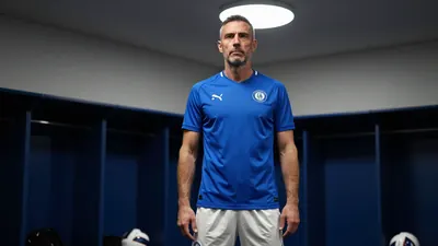 A photorealistic image of an anonymous KV Mechelen left back coach, aged 35-45, with a serious expression, standing in a...