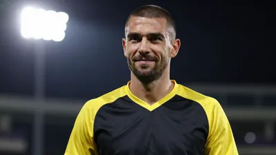 A 25-30 year old male coach with a rugged face and a buzz cut, wearing a black and yellow jersey with a smooth, unbroken...