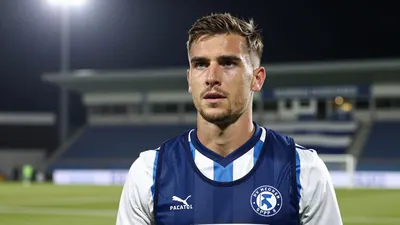 A 25-year-old male footballer in a blue and white striped jersey, standing on a football pitch with a determined...